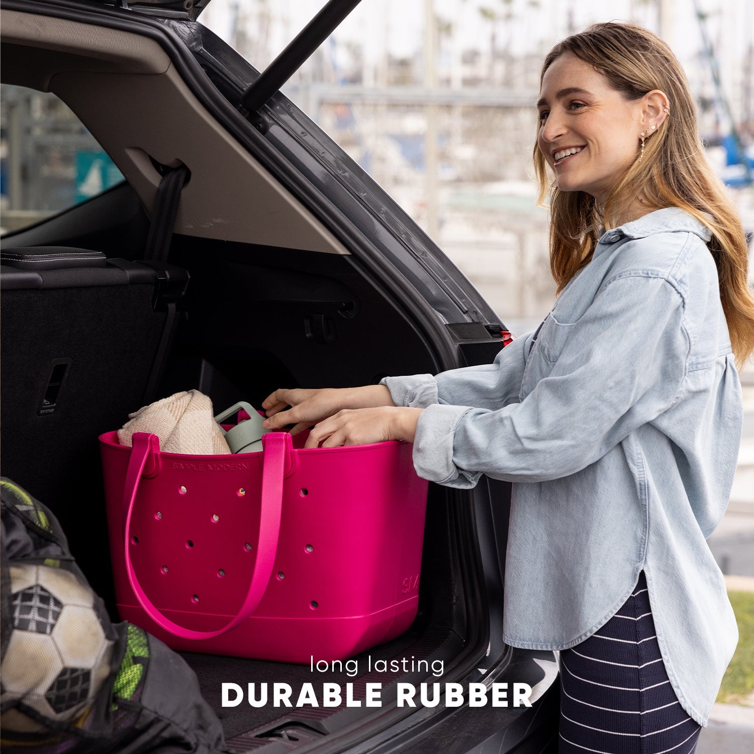 Woman loading a bright hot pink durable rubber tote bag into a car trunk in an urban setting. Bag features ventilation holes and sturdy handles. Text reads "long lasting" and "DURABLE RUBBER."