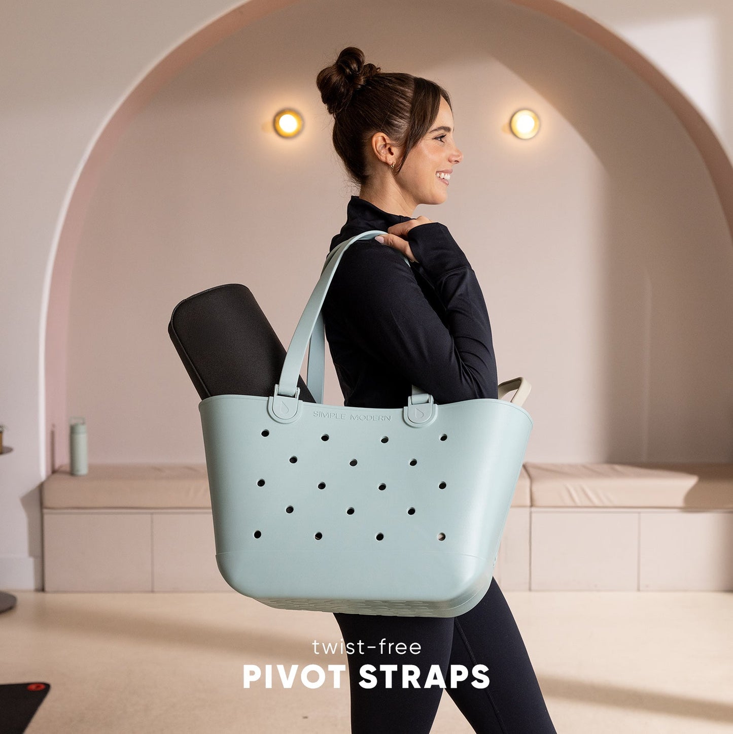 Woman in black athletic wear holding a light mint blue tote bag with black perforations and twist-free pivot straps in a modern studio with curved archway backdrop.