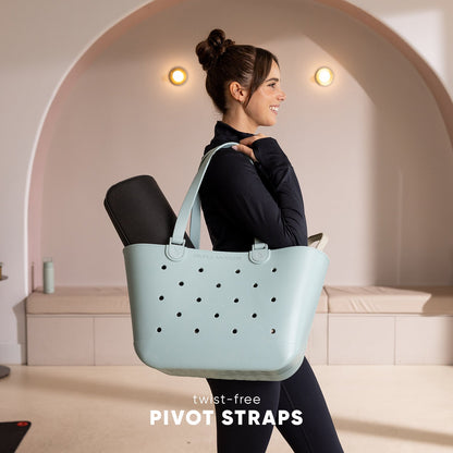 Woman in black athletic wear holding a light mint blue tote bag with black perforations and twist-free pivot straps in a modern studio with curved archway backdrop.