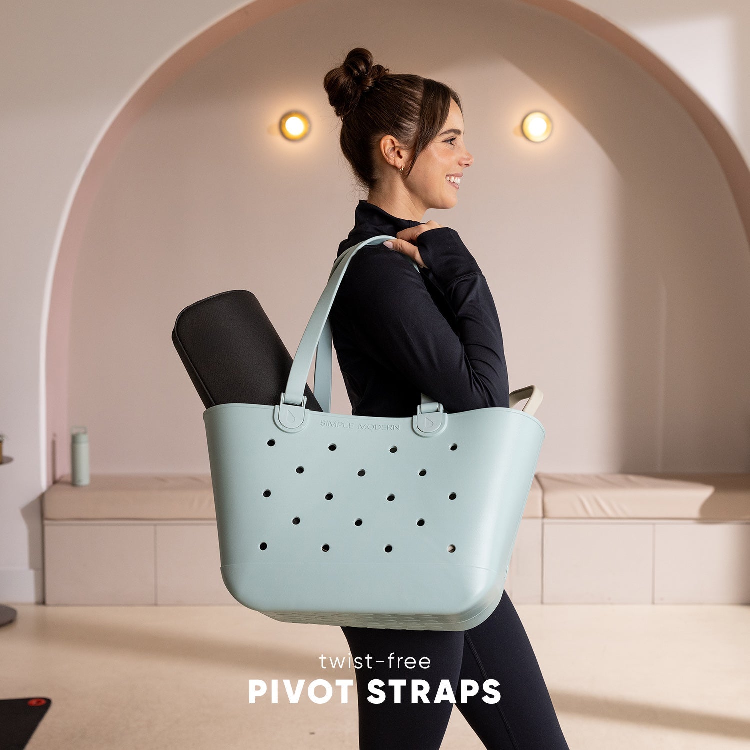 Woman wearing black athletic top and leggings carries a pale blue-green perforated tote bag with twist-free pivot straps over her shoulders in a modern studio setting with curved architecture.