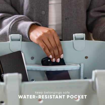 Person placing black smartphone into water-resistant pocket of light green Simple Modern Getaway Bag, demonstrating pocket functionality and storage capacity for keeping belongings safe.