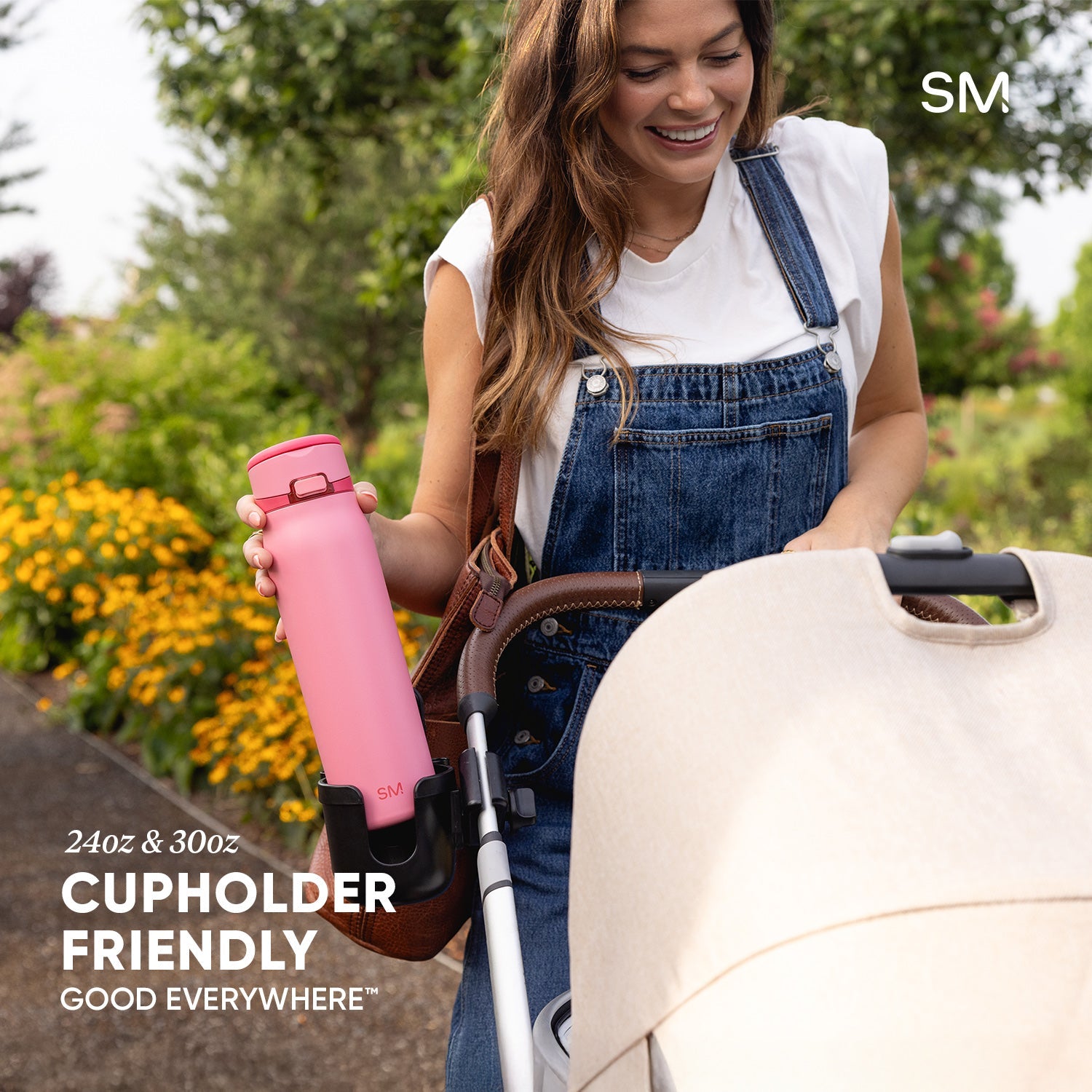 Woman in overalls holding a bright pink SM water bottle in a garden with a cream-colored stroller featuring a cupholder attachment; text reads "24oz & 30oz," "Cupholder Friendly," and "Good Everywhere."