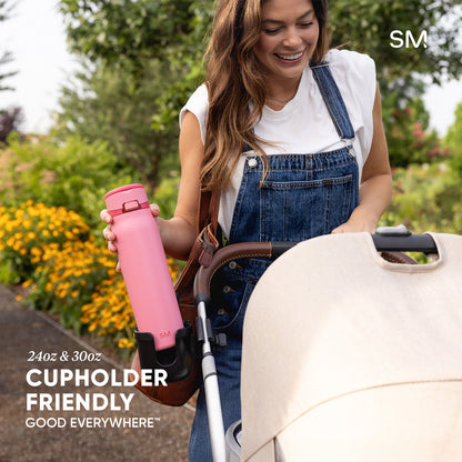 Woman in overalls holding a bright pink SM water bottle in a garden with a cream-colored stroller featuring a cupholder attachment; text reads "24oz & 30oz," "Cupholder Friendly," and "Good Everywhere."