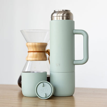 Sage green insulated thermos next to matching cup and Chemex coffee brewer with wood-tone band, on white background